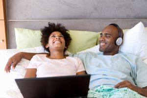 Young couple sitting on bed with laptop, man wearing headphones, smiling