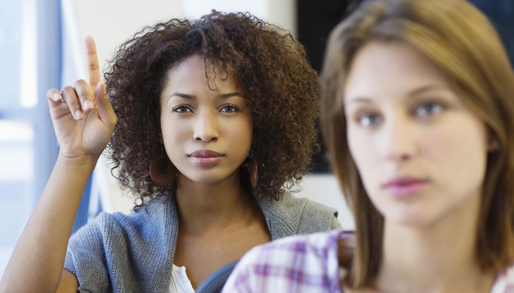 Two women sitting in classroom with focus on African American woman raising hand