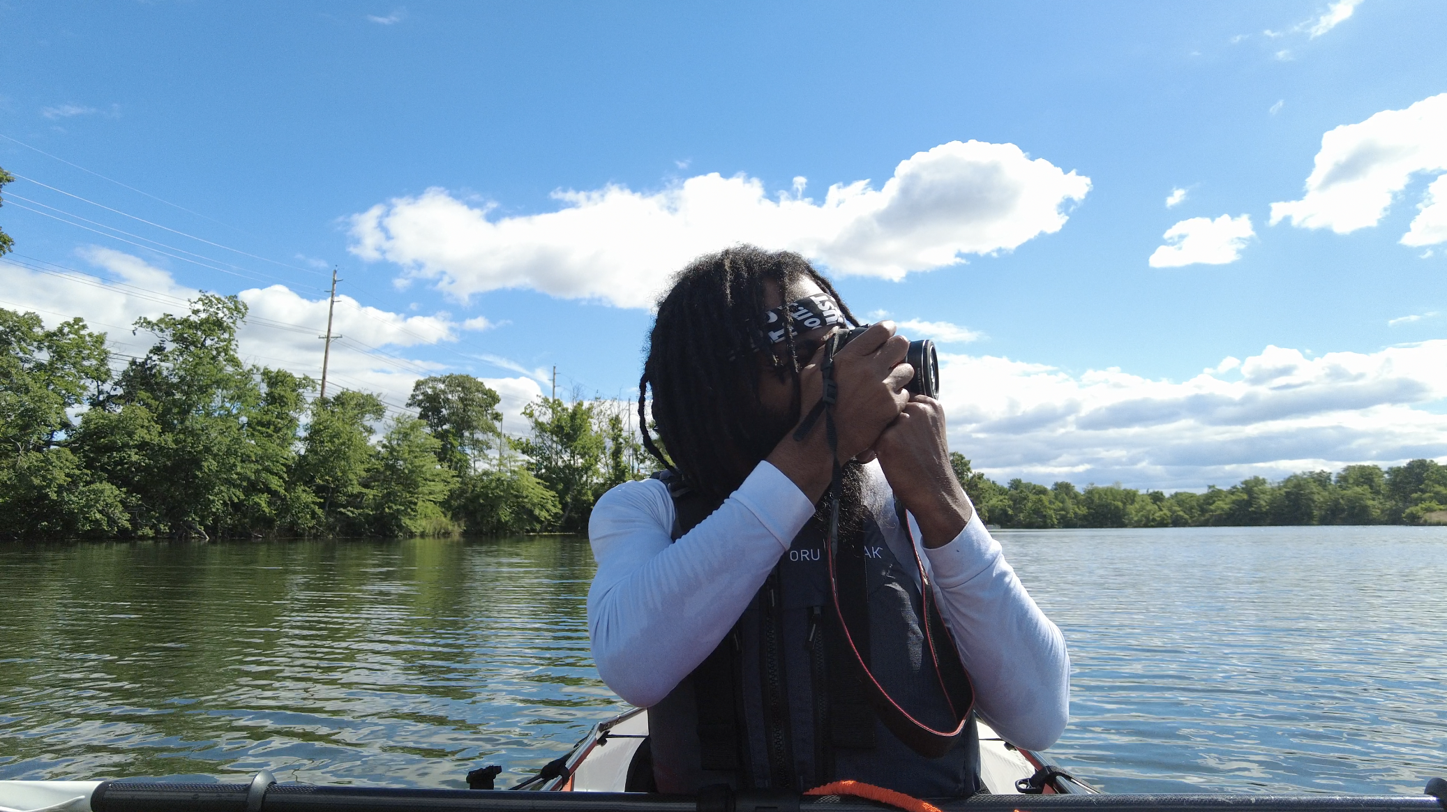 A person wearing a white shirt and black jacket is taking a photograph while sitting in a boat on a lake surrounded by trees and a cloudy sky.