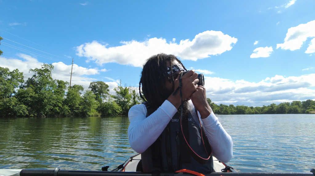 A person wearing a white shirt and black jacket is taking a photograph while sitting in a boat on a lake surrounded by trees and a cloudy sky.