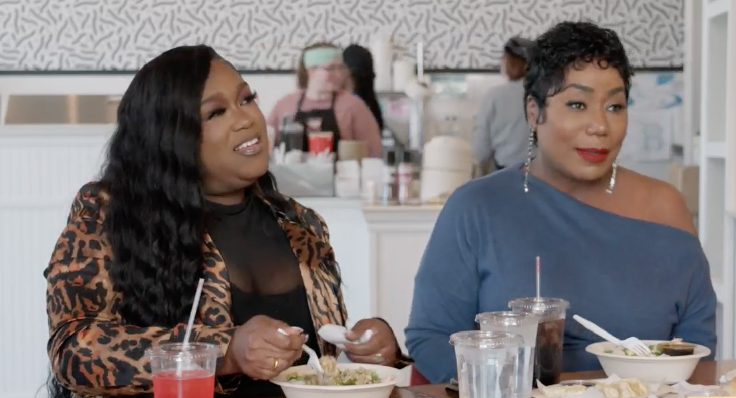 Two women sitting at a table, eating and drinking, in what appears to be a restaurant or cafe setting. One woman is wearing a leopard print jacket, the other a blue top.