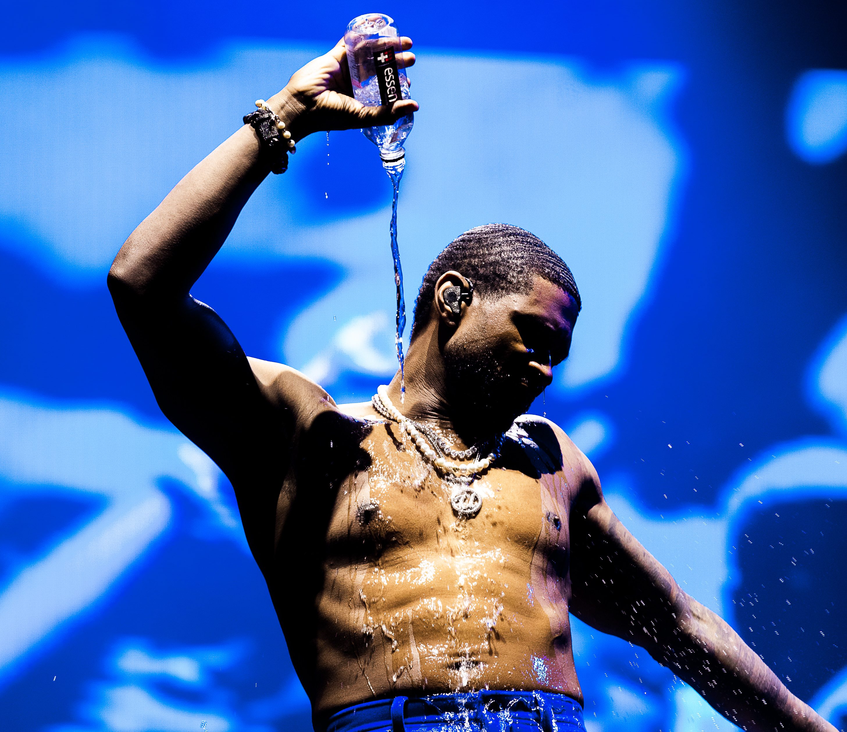 A shirtless man holding a water bottle and standing in front of a blue background, with water splashing down his body.