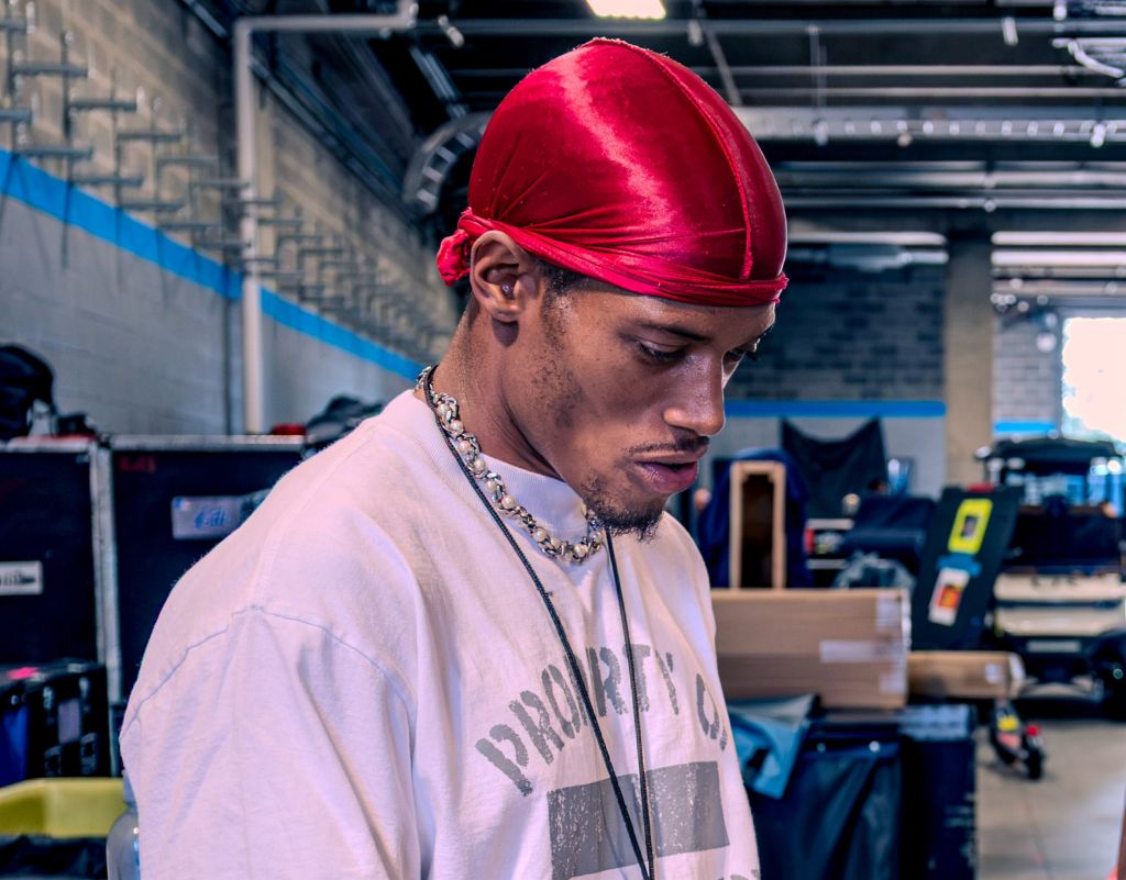 A young man wearing a red headband and a white shirt with "Droped" printed on it, standing in a room with various equipment.