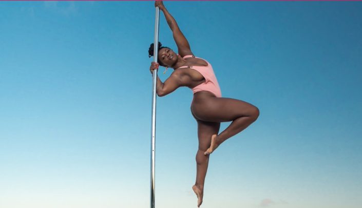 A person in a pink swimsuit performing an acrobatic pole dance move over the ocean with the text "Fitness Fridays" visible.