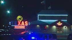 Nighttime scene of a Xas convenience store with police car and other vehicles parked outside.