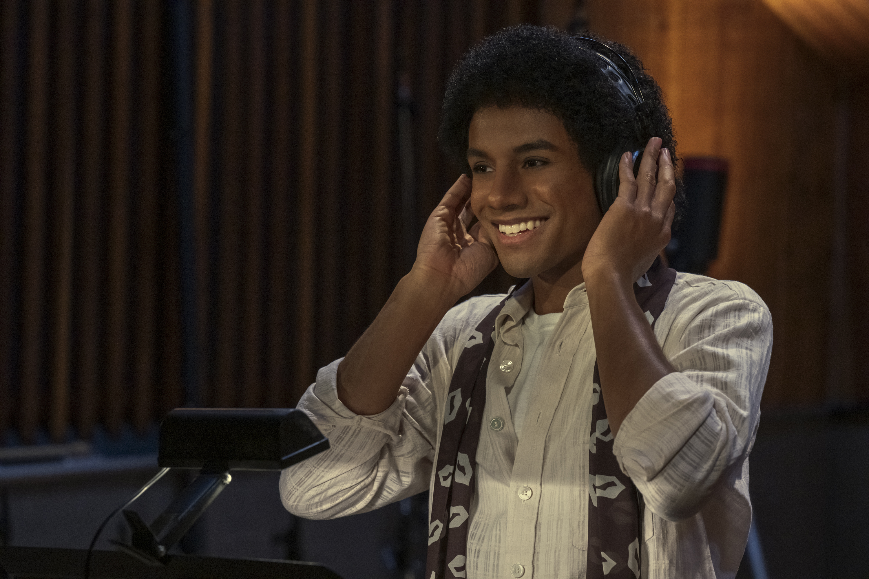 A smiling young man wearing a white shirt and scarf, listening to headphones and performing on a stage.