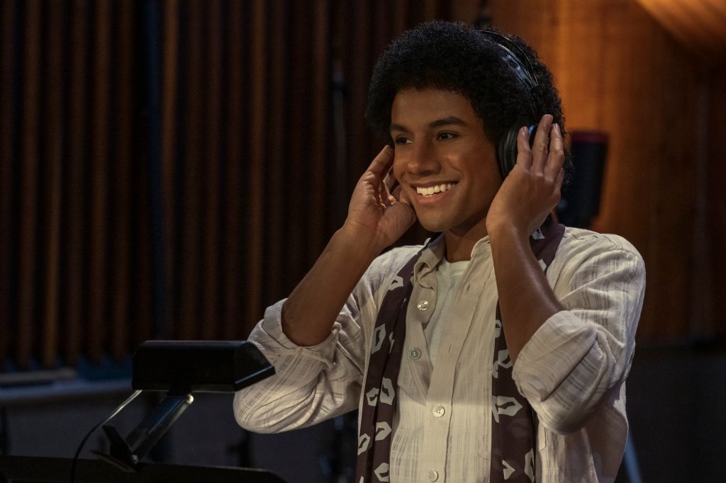 A smiling young man wearing a white shirt and scarf, listening to headphones and performing on a stage.