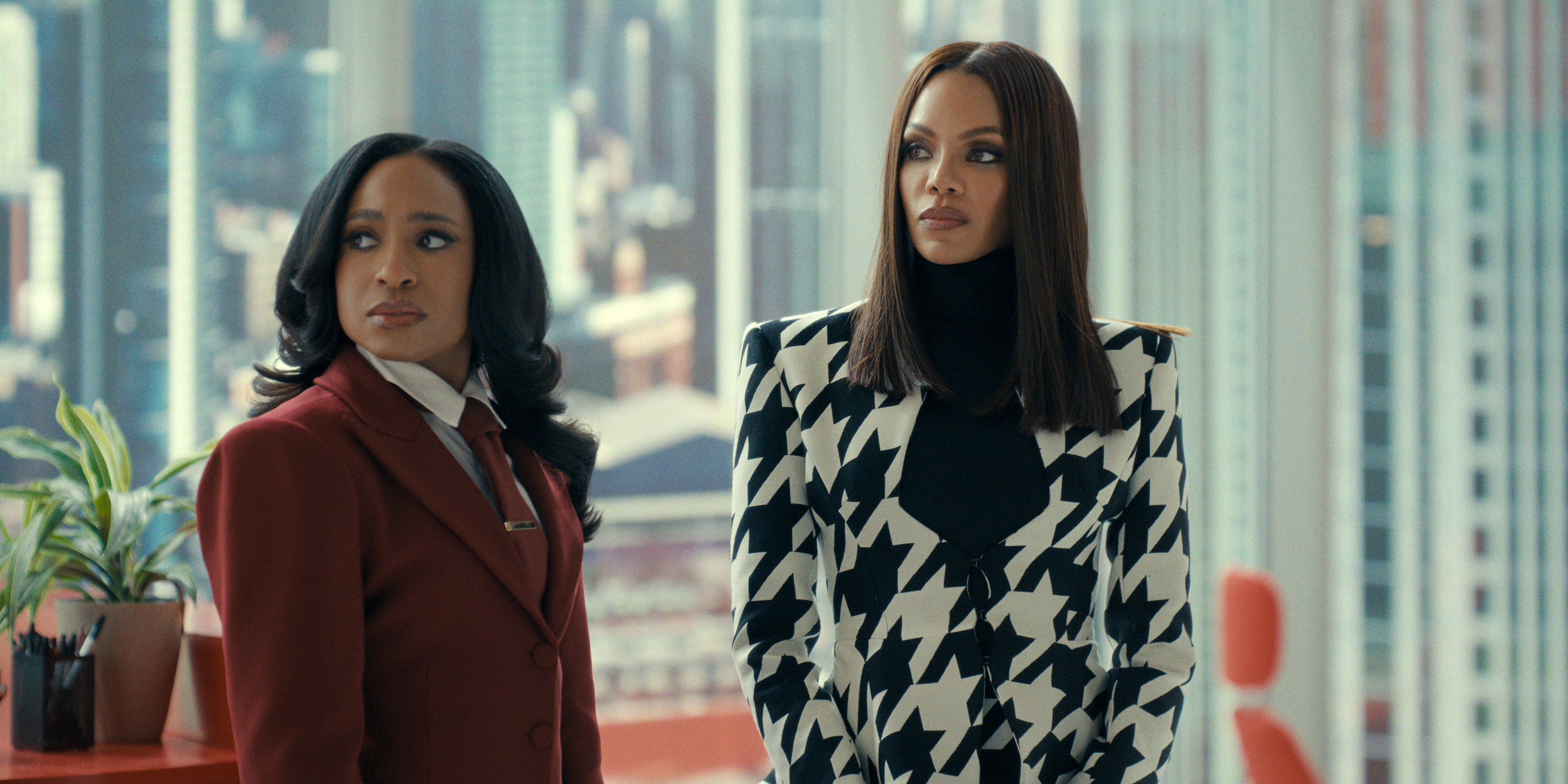 Two women, one in a red coat and the other in a black and white patterned coat, standing in an urban setting with buildings in the background.